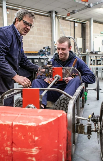 A lecturer assisting a student seated in a custom-built electric car, adjusting the wiring and components during a hands-on engineering project. A lecturer assisting a student seated in a custom-built electric car, adjusting the wiring and components during a hands-on engineering project.